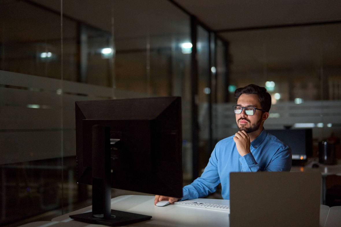 A software developer wearing glasses sits at a desk, focused on his computer screen while working on a project.