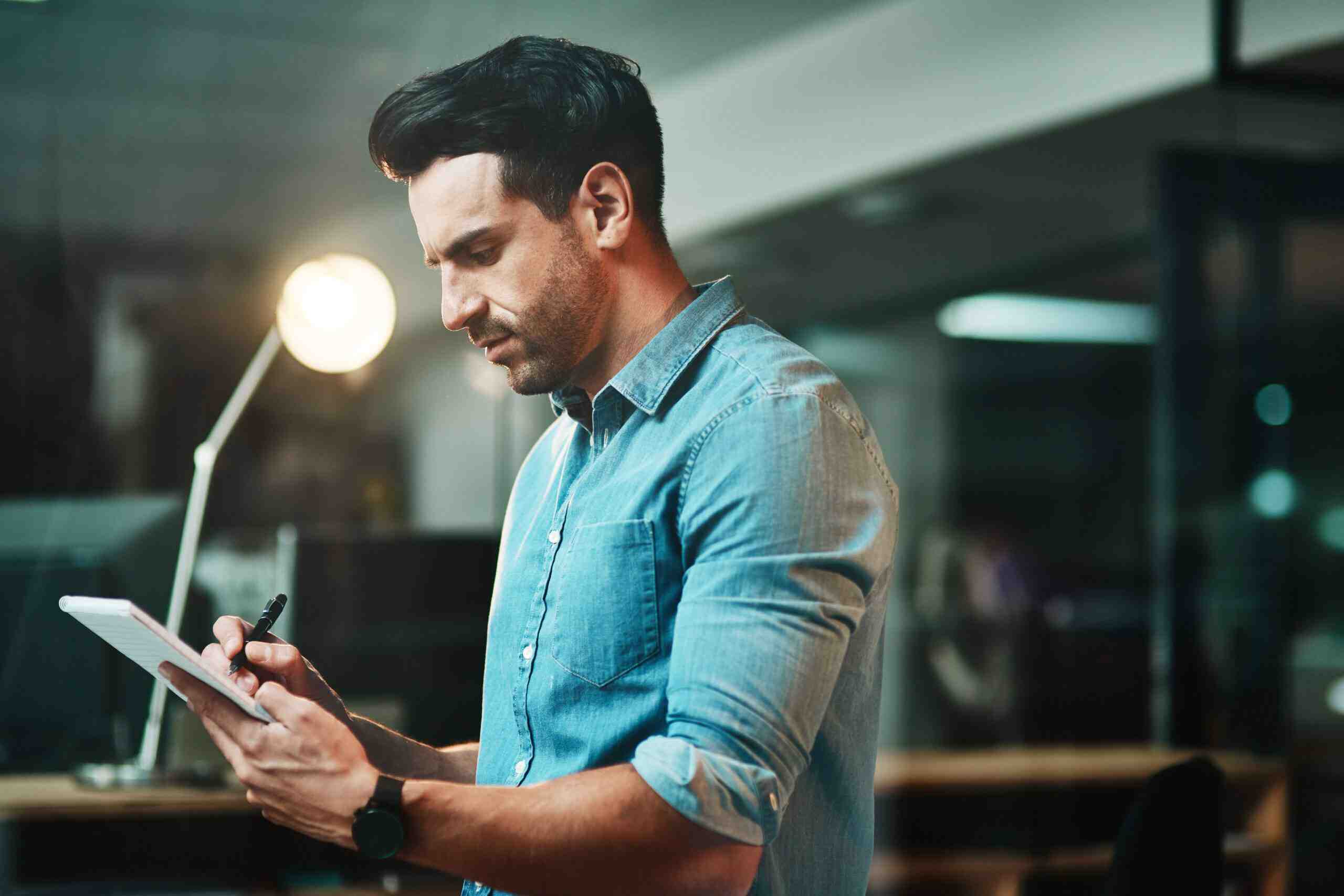 A man focused on writing on a tablet while seated at a desk in a modern office environment.