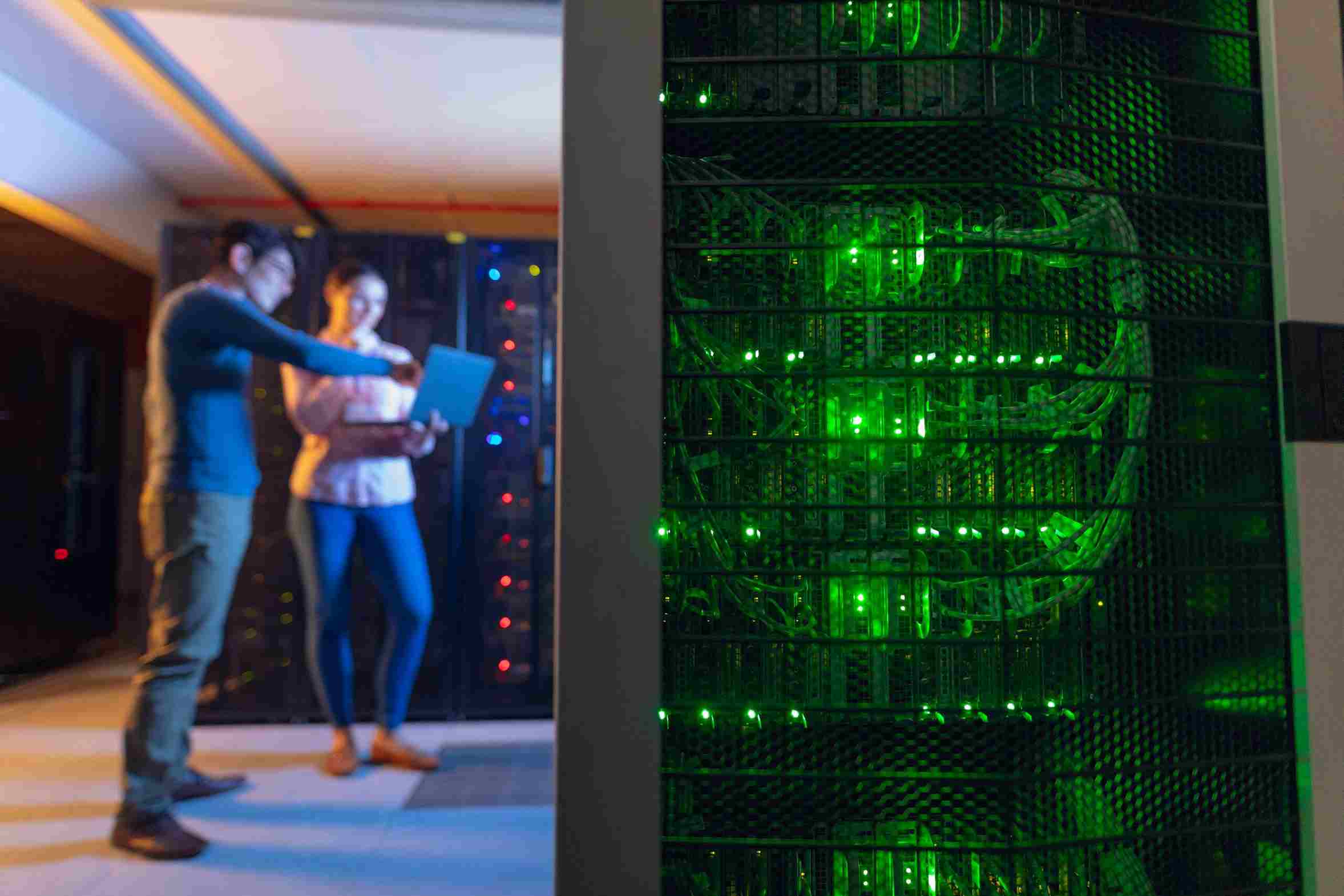 Two people stand in a dimly lit room, working on laptops in front of a large server rack with green lights. The server's intricate wiring is visible.