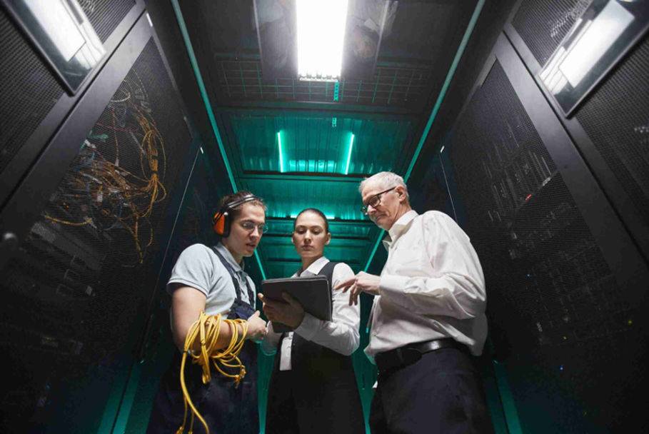 Three individuals in a server room examining a tablet, surrounded by servers and technology equipment.