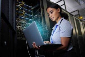 Laptop, network and data center with a black woman it support engineer working in a dark server roo.