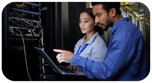 Two TRT employees collaborating on a laptop in a server room, focused on solving a technical issue together.